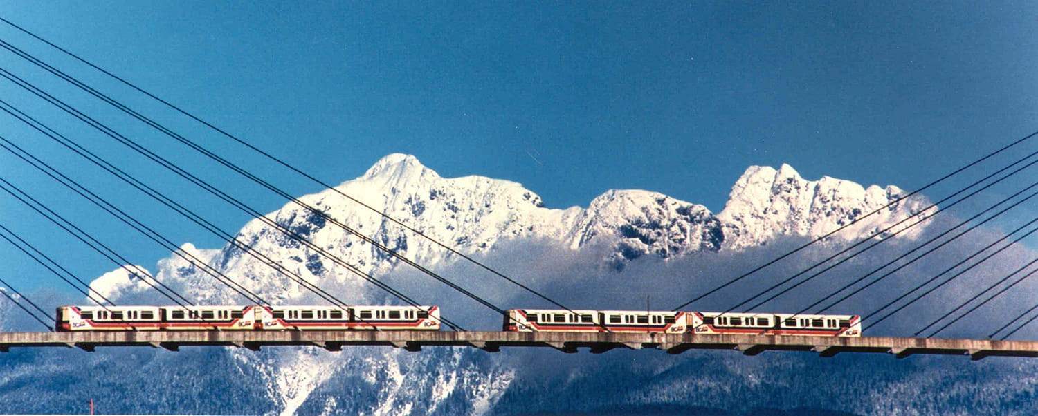 A Mark 1 SkyTrain running on a bridge with snowy mountains and a clear sky in the background