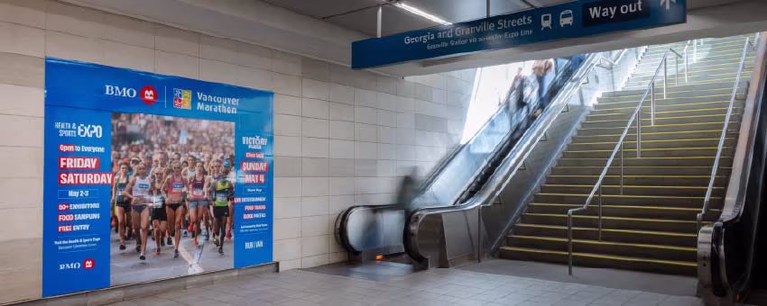 A large poster advertisement for the 2025 BMO Vancouver Marathon is on display at the Vancouver-City Centre Station concourse on the Canada Line. There are stairs and escalators leading riders up to the intersection of West Georgia and Granville Street.