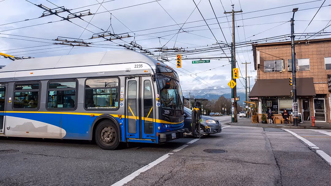 A TransLink trolleybus passing through an intersection, with overhead wires, traffic lights, and nearby shops visible in the background.