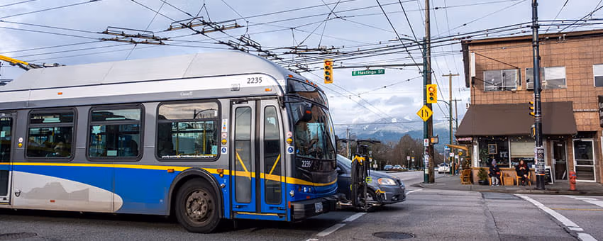 A TransLink trolleybus passing through an intersection, with overhead wires, traffic lights, and nearby shops visible in the background.