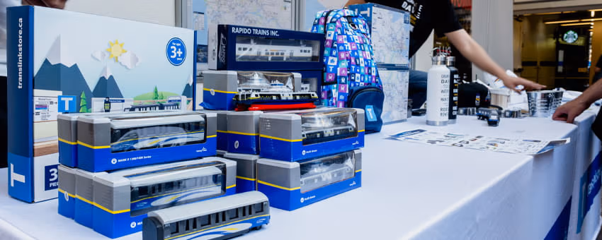 A display table showcases various TransLink store merchandise, including trains, a SeaBus, a backpack, a water bottle, and other souvenirs.