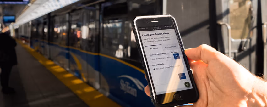 Mobile phone on ‘Transit Alerts’ page held up in front of SkyTrain at station platform