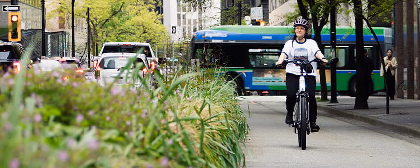 A person riding a bicycle on a city bike path, wearing a helmet and a white T-shirt, with the Translink RapidBus bus and cars in the background.