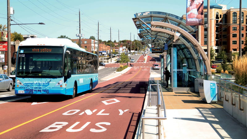 A conceptual illustration of a Bus Rapid Transit (BRT) system showing a modern bus in a designated bus lane approaching a station with waiting passengers.