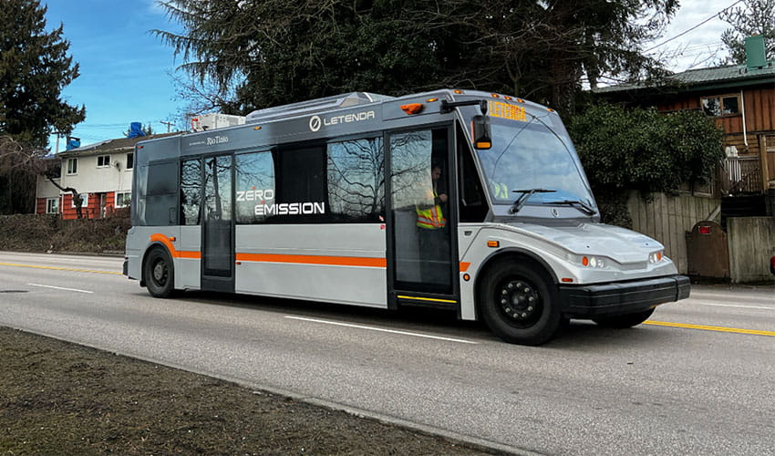 A compact, silver Letenda zero-emission battery- electric bus with black and orange accents is driving on a suburban street.