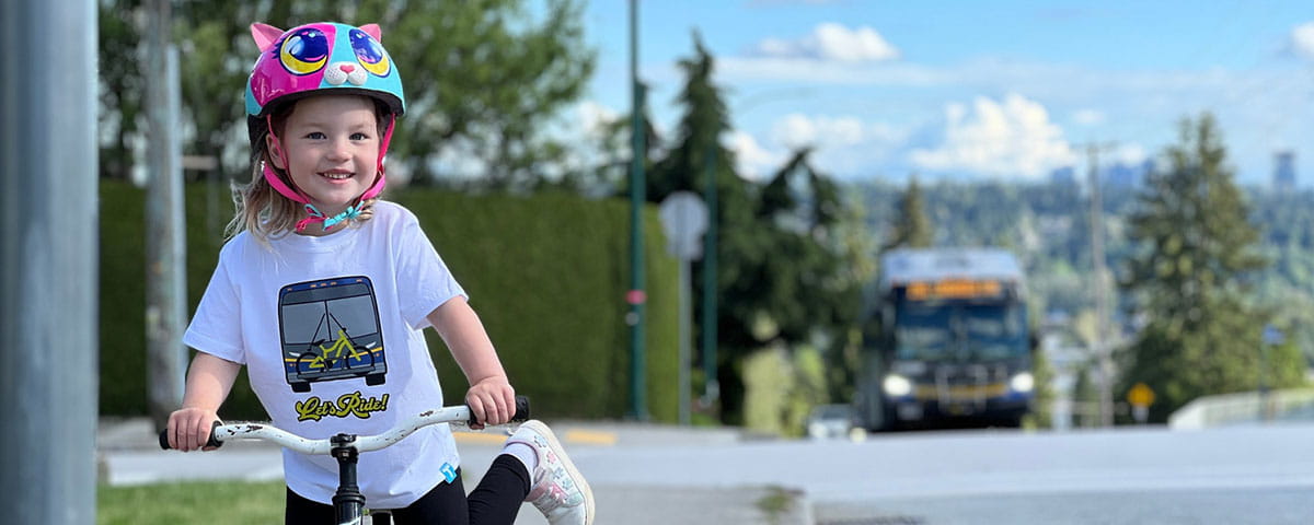 A young girl wearing a helmet and riding her bicycle. In the background, a city bus is approaching on the street.