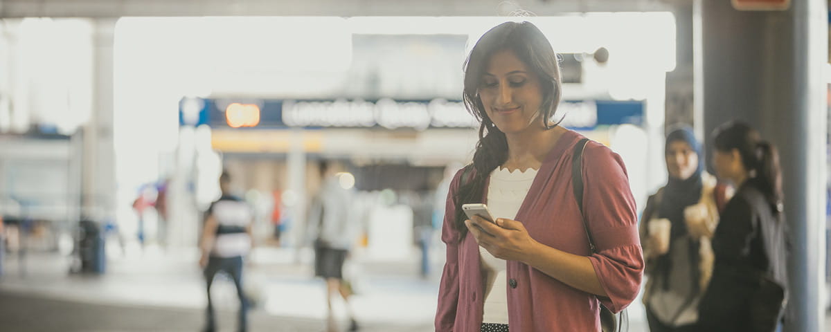 A woman standing at a transit station, smiling as she looks at her smartphone.