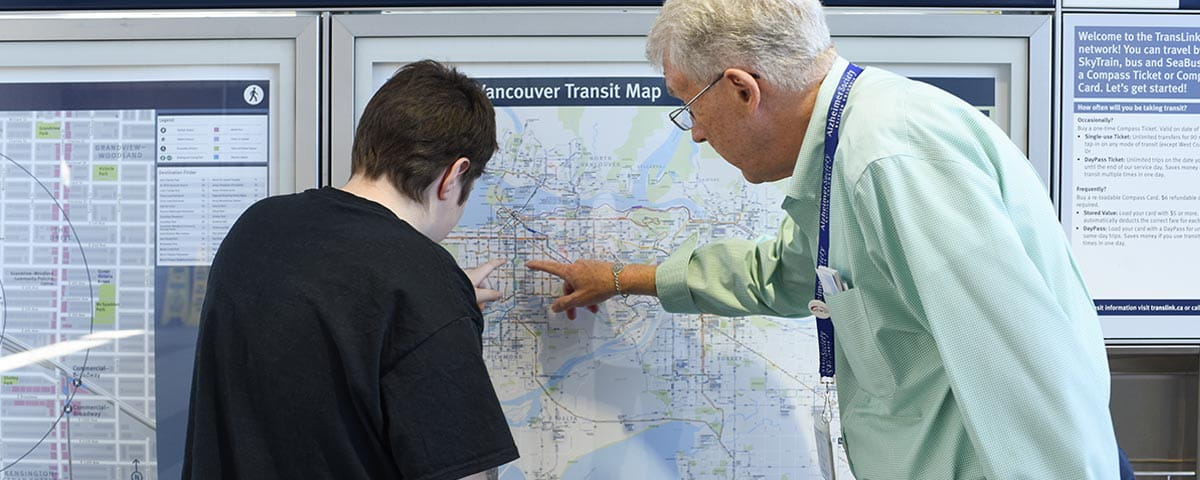 A senior assists a young passenger by pointing out directions on a detailed Vancouver transit map