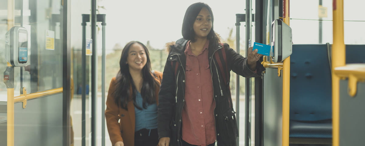 A girl taps her student Compass Card before entering a bus, while her friend waits behind her.