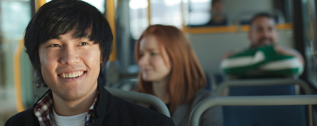 A young man with black hair and a friendly smile sits on a bus, with other passengers visible in the background.