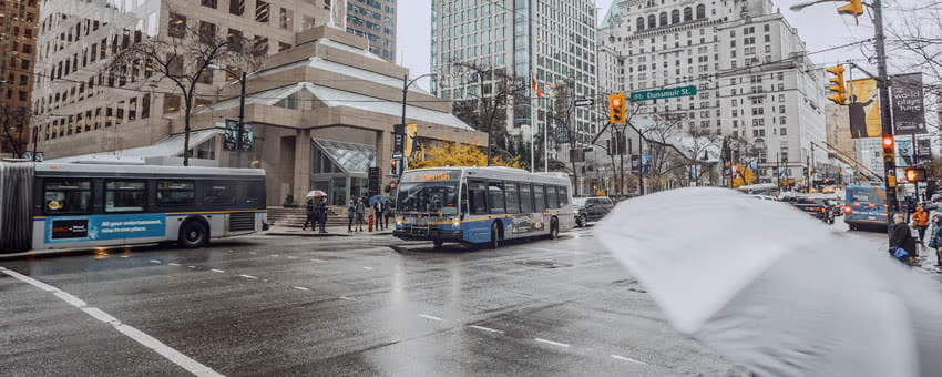 Two buses downtown during rainy weather with a grey umbrella in the foreground