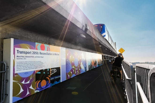 View of Canada Line Bridge Indigenous murals with cyclist looking at them and SkyTrain passing overheard.