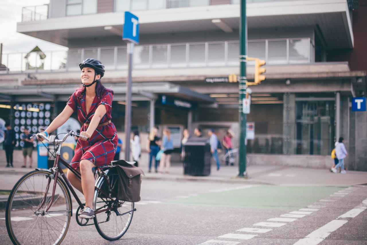 A woman is crossing the street while riding her bicycle, a SkyTrain station is in the background