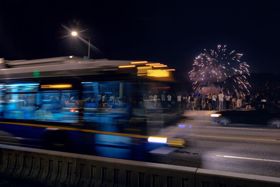 Blurry bus driving at night on Burrard Street Bridge with pedestrians watching fireworks in background