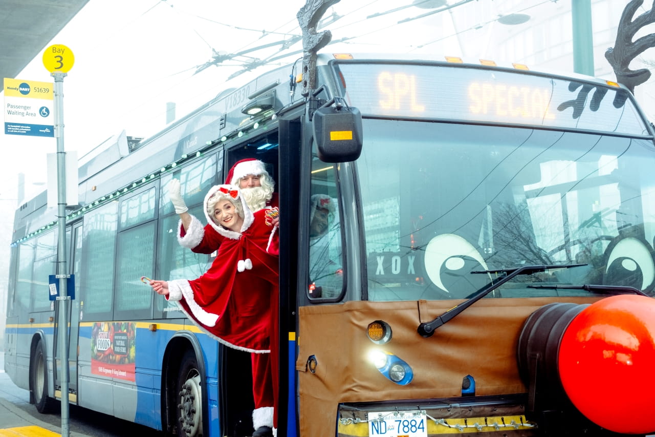 Santa and Mrs. Claus are waving from inside of a fully decorated Reindeer Bus, complete with antlers, eyes, and a red nose